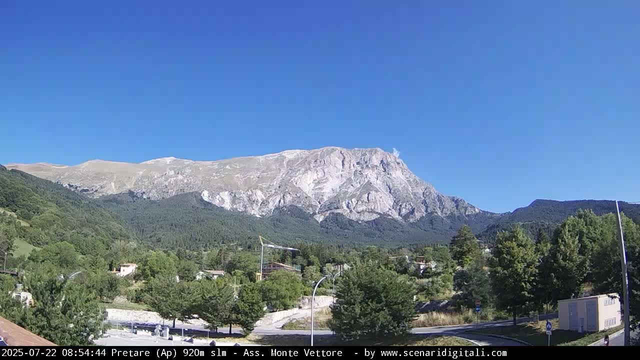 A large, light-colored mountain with some snow is visible in the distance under a clear blue sky, behind a valley of green trees and a small town.