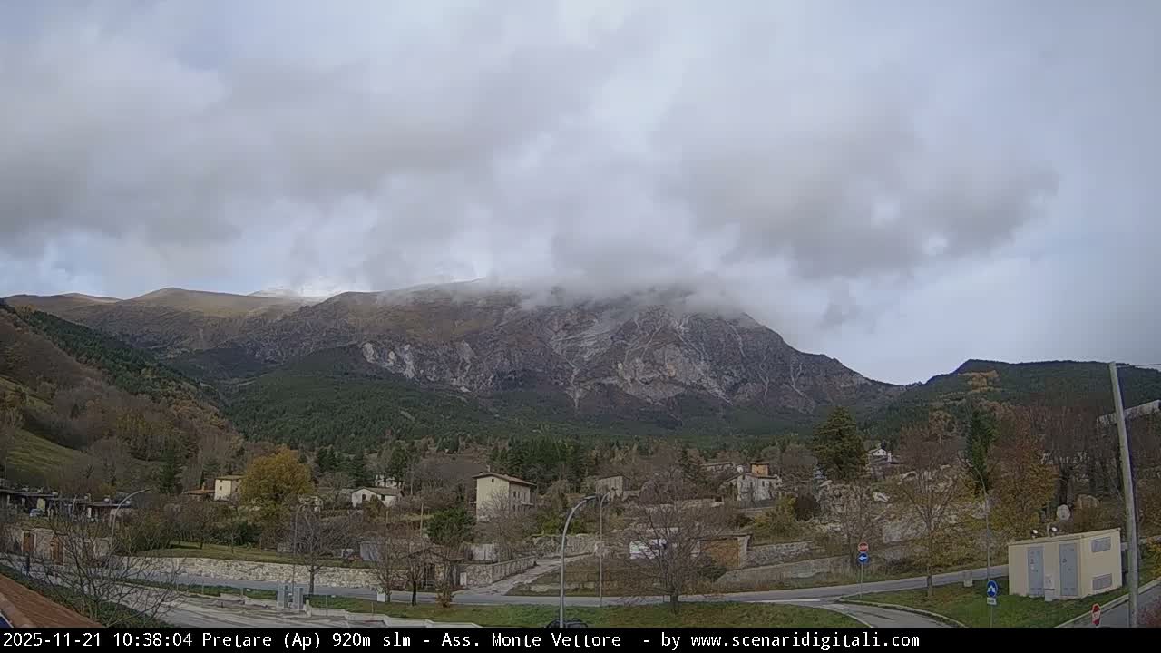 A rugged mountain range shrouded in low, thick clouds overlooks a valley containing a scattered village, roads, and a mix of green and bare trees under an overcast sky.