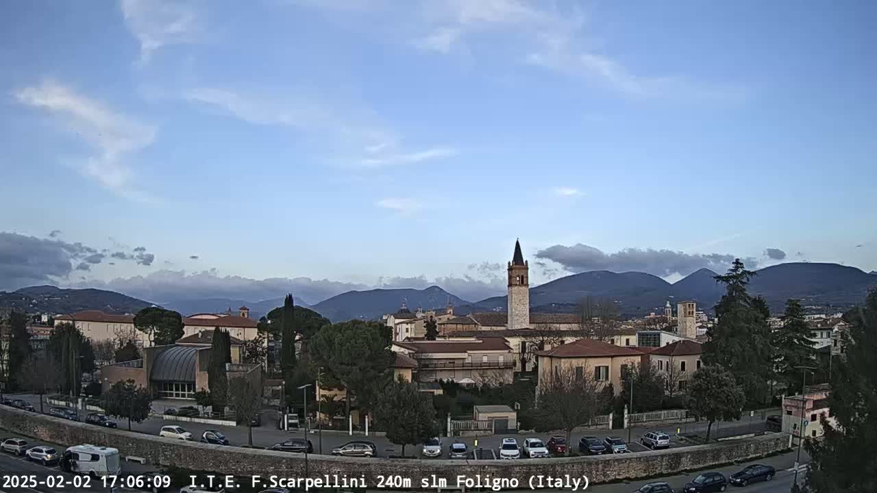 A partly cloudy day reveals a view of a town nestled in a valley, with mountains in the background and a tall bell tower prominently featured among the buildings.