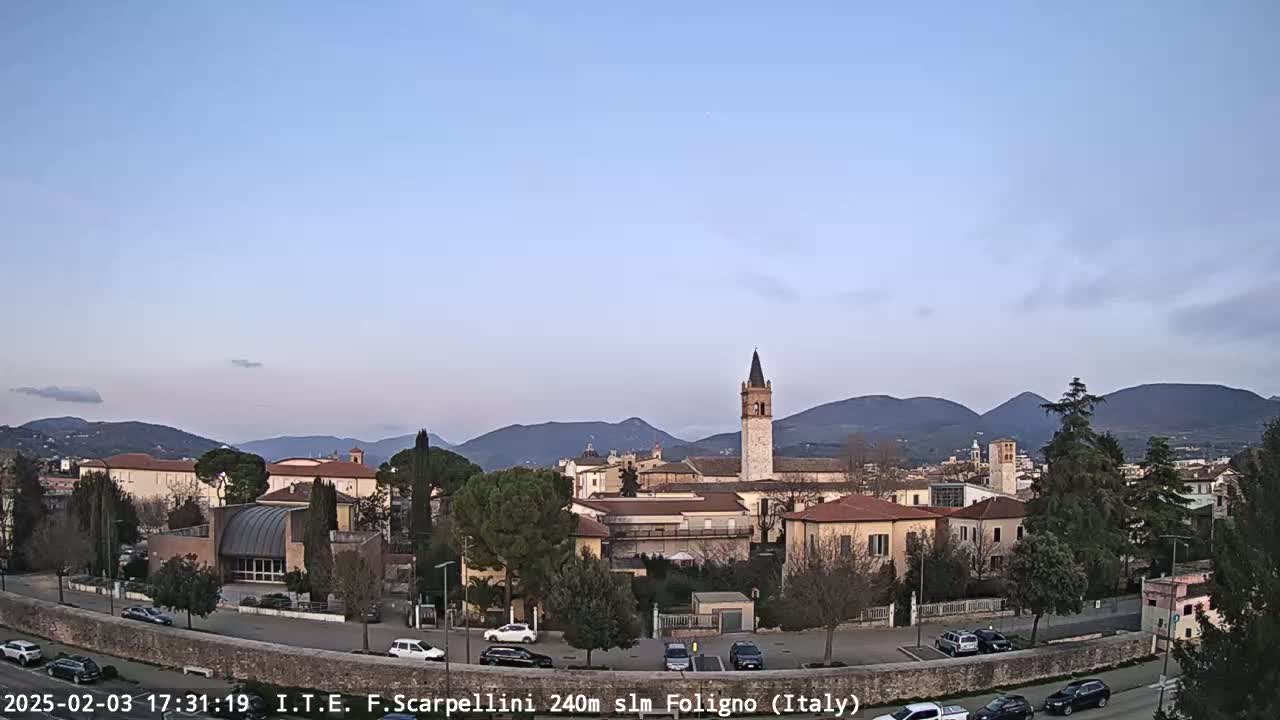A clear, twilight view shows a town nestled in a valley, with a tall bell tower prominently featured, and mountains in the background.