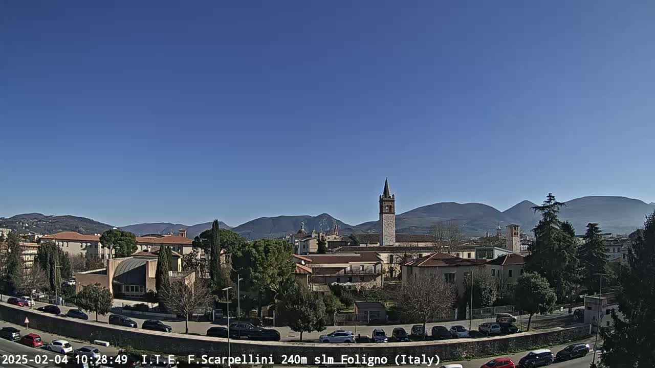 A sunny day reveals a town nestled in a valley, with mountains in the background and a tall bell tower prominent among the buildings.