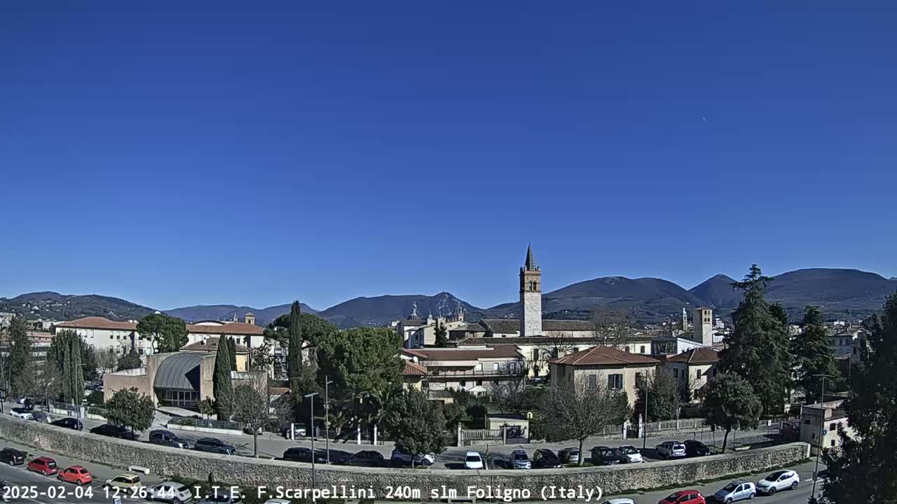 A clear, sunny day reveals a picturesque town nestled in a valley, with mountains in the background and cars parked along a road in the foreground.