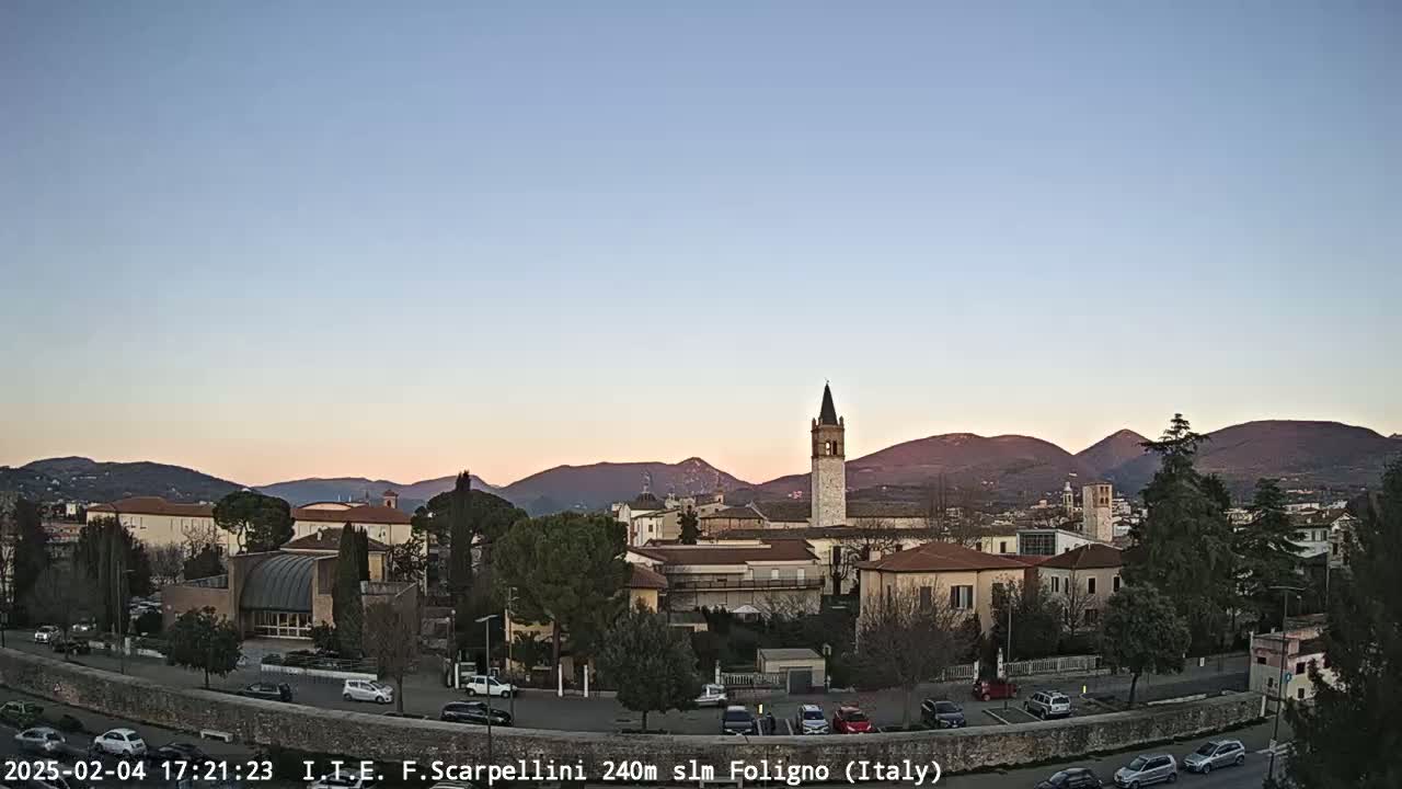 A town with a tall bell tower is nestled in a valley at sunset, with mountains in the background under a clear sky.