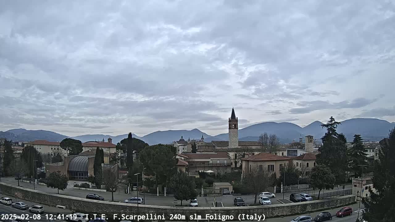 A mostly cloudy sky overlooks a town with low buildings, a tall bell tower, and mountains in the distance.
