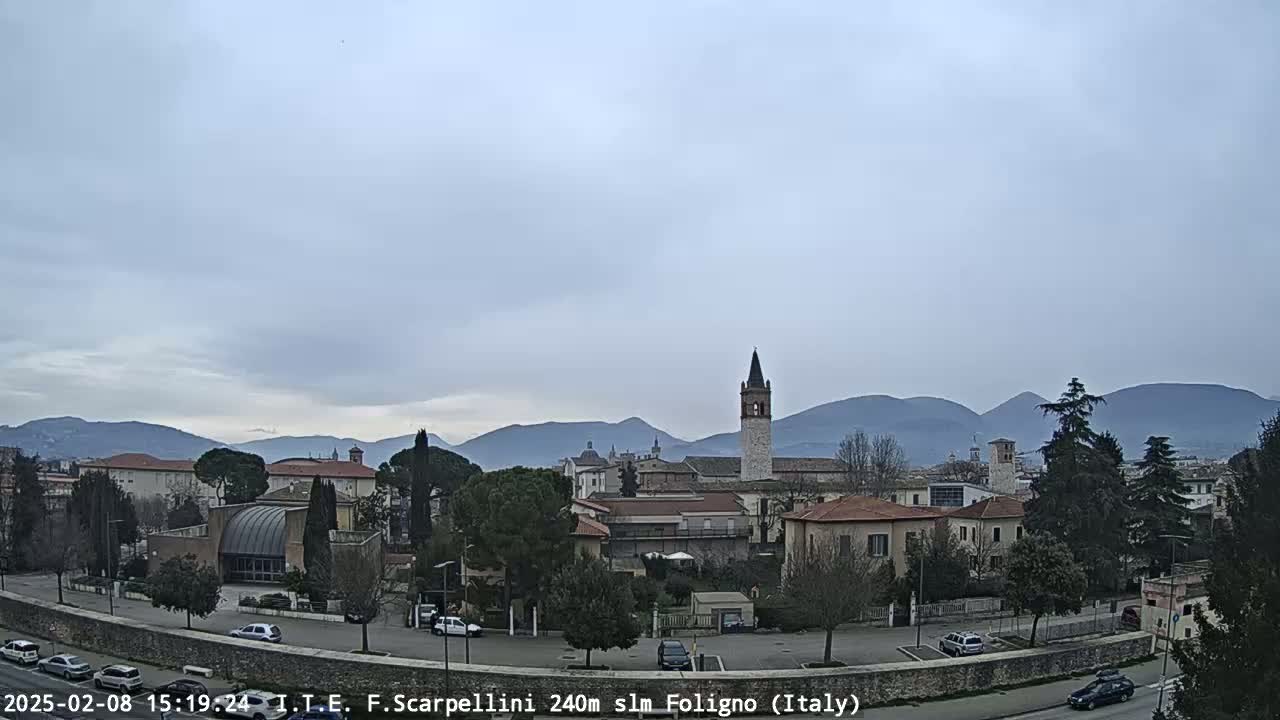 A town with several buildings and a tall bell tower is nestled in a valley, with mountains in the background under an overcast sky.