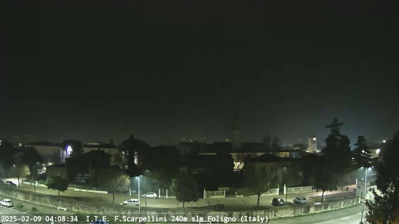 A nighttime view of a town under a hazy sky shows a few cars parked and driving on roads that curve through the area.
