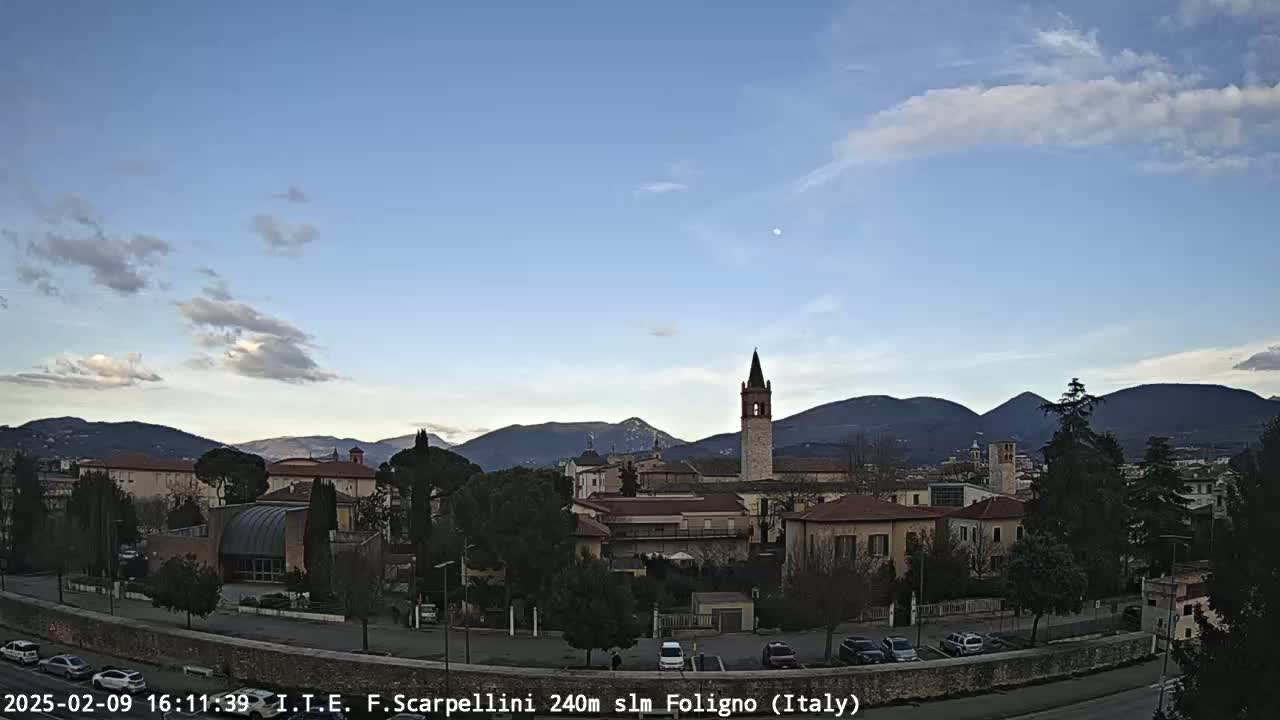 A partly cloudy afternoon shows a town nestled in a valley between mountains, with a tall bell tower prominently featured in the center.