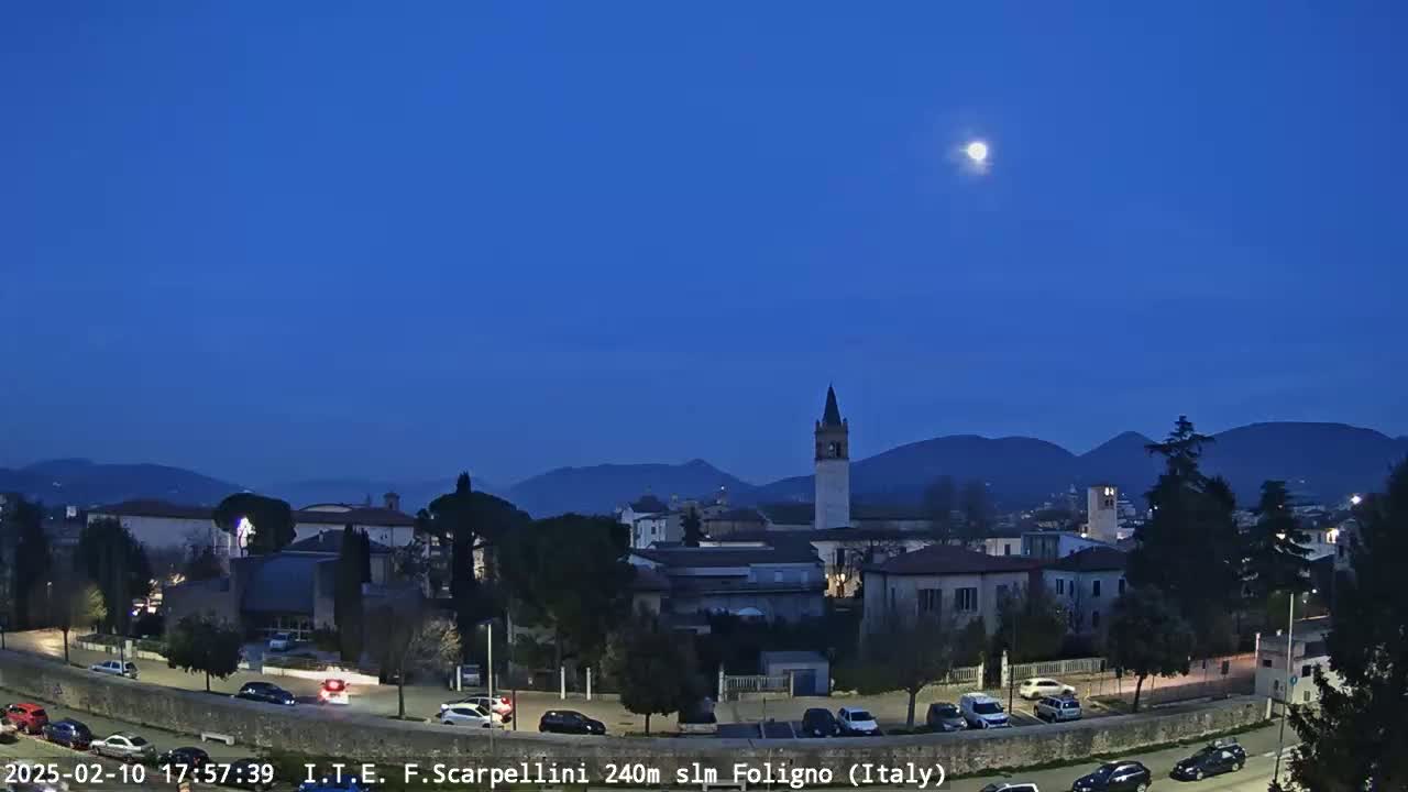 A moonlit night scene shows a town nestled in a valley, with mountains in the background and cars parked along a road in the foreground.