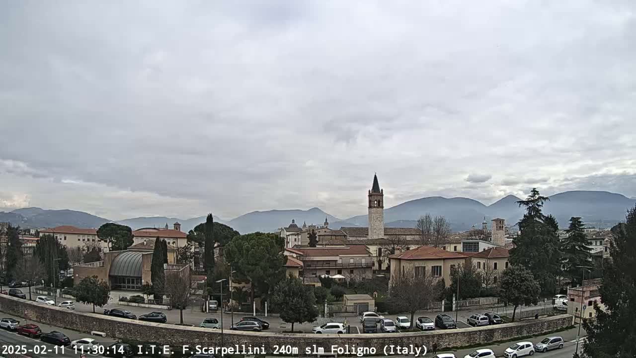 A view of a town with buildings and a tall clock tower, under an overcast sky with mountains in the background.