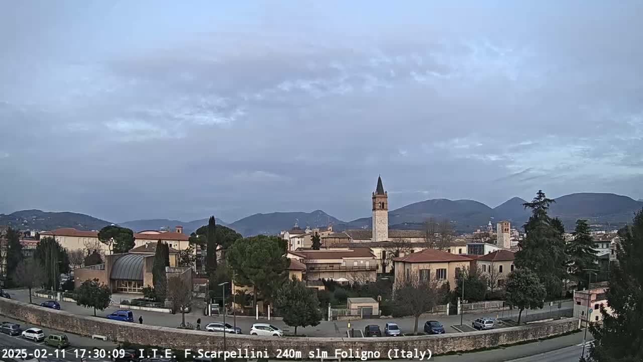 A town with low buildings and a tall tower is situated in a valley under a cloudy sky, with mountains visible in the background.