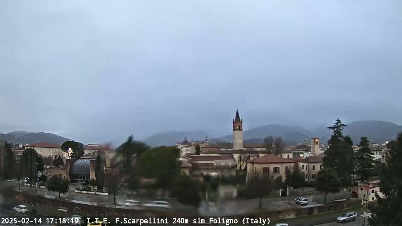 A rainy view of a town in a valley, with buildings, trees, and a tall tower visible under a cloudy sky.