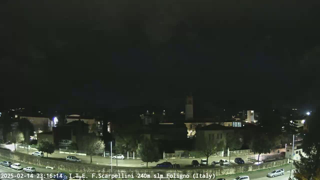 A nighttime view of a town with buildings, trees, and parked cars under a dark, cloudy sky.