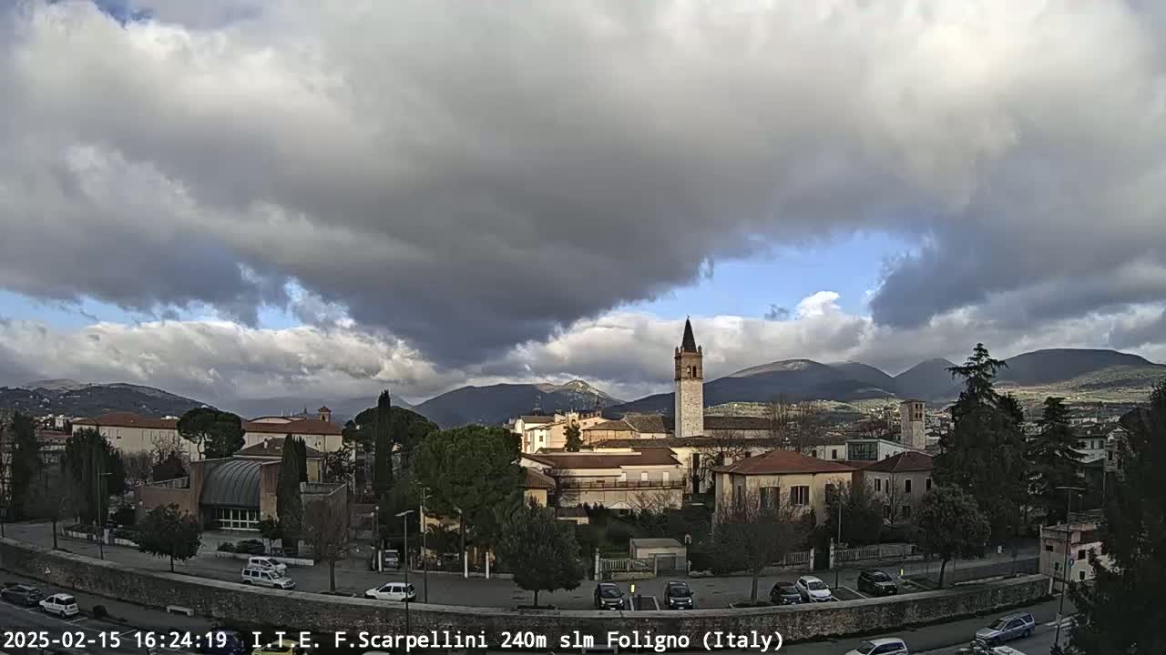 A partly cloudy day reveals a town nestled in a valley between mountains, with a tall church steeple prominent in the center.