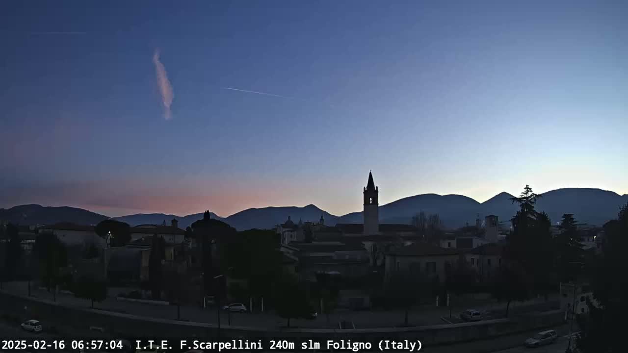 A twilight view shows a town nestled at the base of silhouetted mountains under a clear, dark-blue sky with a single, thin, vertical cloud and a contrail.