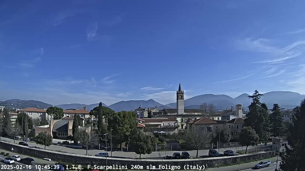 A clear, sunny day reveals a cityscape nestled amongst mountains, featuring buildings with terracotta roofs and a prominent bell tower.