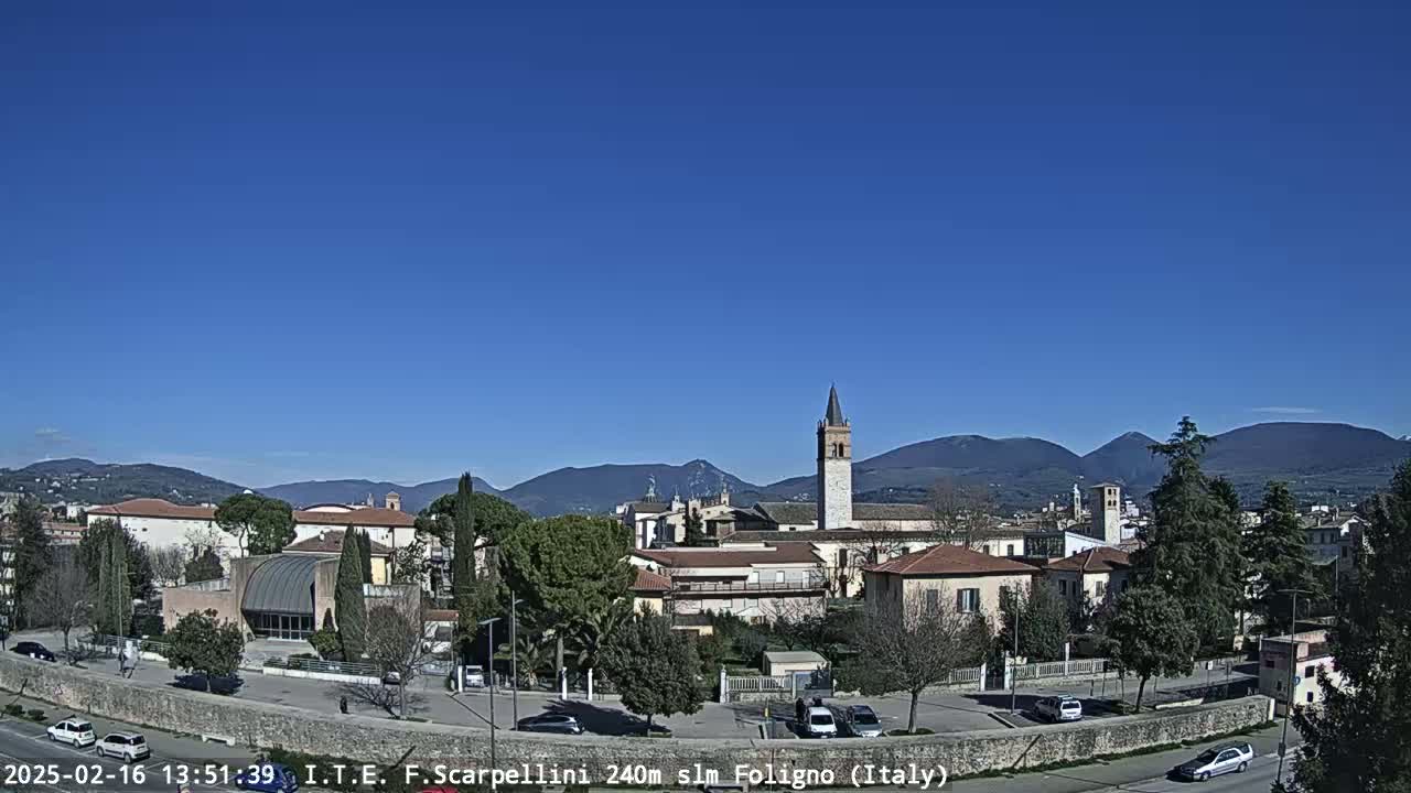 Under a clear blue sky, a town with low buildings, a tall bell tower, and some trees sits before a range of mountains in the distance.