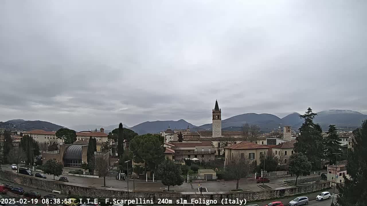 A town with low buildings and a tall bell tower is situated in a valley, with mountains in the background under an overcast sky.