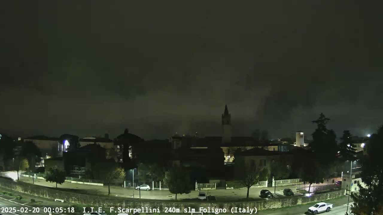 A nighttime view of a town under a cloudy, dark sky shows buildings, trees, and a few parked cars along a road.