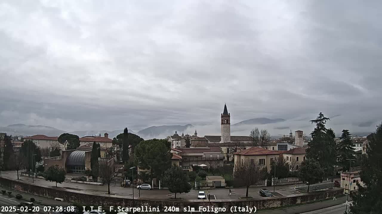 A town with low buildings and a tall bell tower is nestled in a valley under a cloudy sky, with mountains in the distance partially obscured by fog.