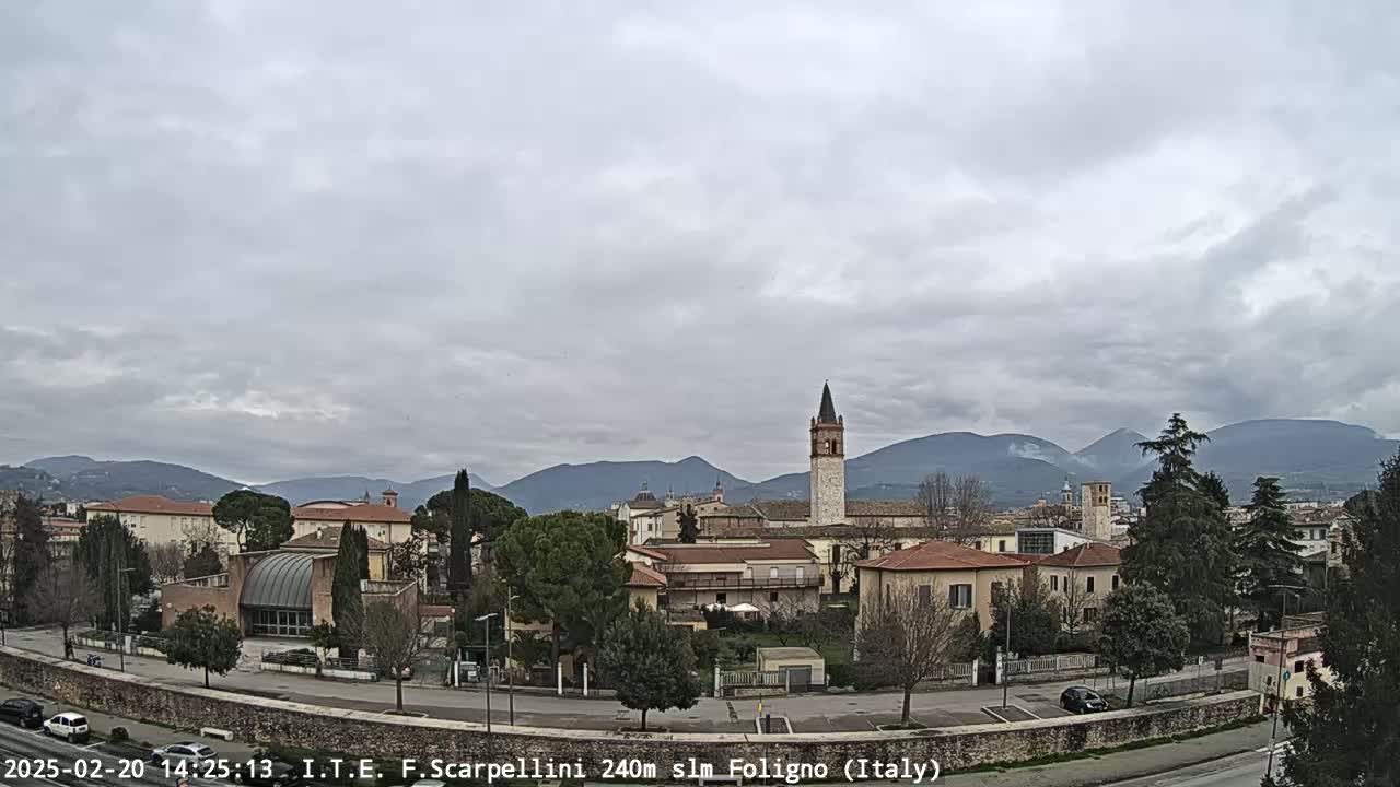 A view of a town nestled in a valley, under a cloudy sky, with mountains visible in the background.
