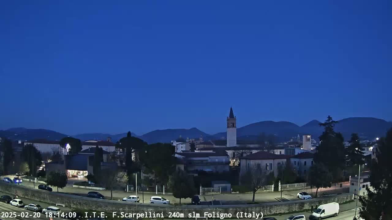 Under a twilight blue sky, a town nestled in a valley is shown with its buildings, a tall bell tower, and distant mountains.