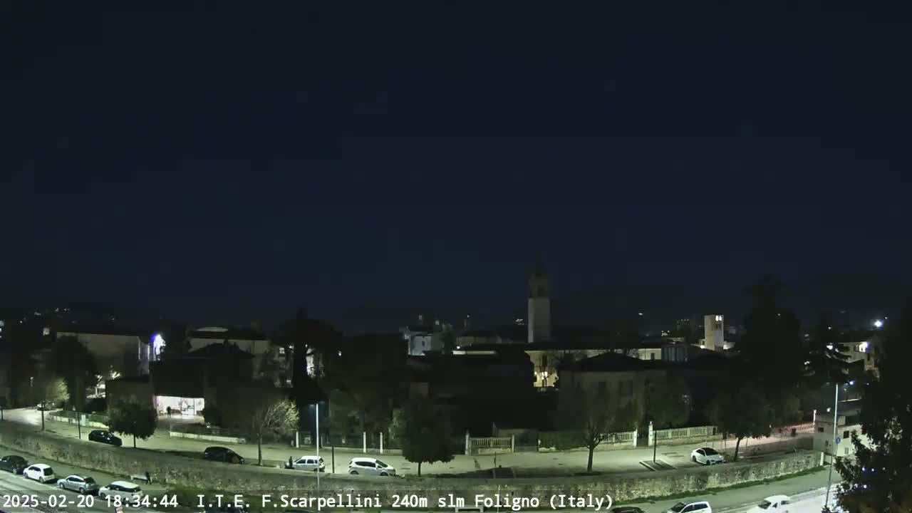 A nighttime, high-angle view of a town with a few cars on roads and a tall tower in the center, under a clear dark sky.