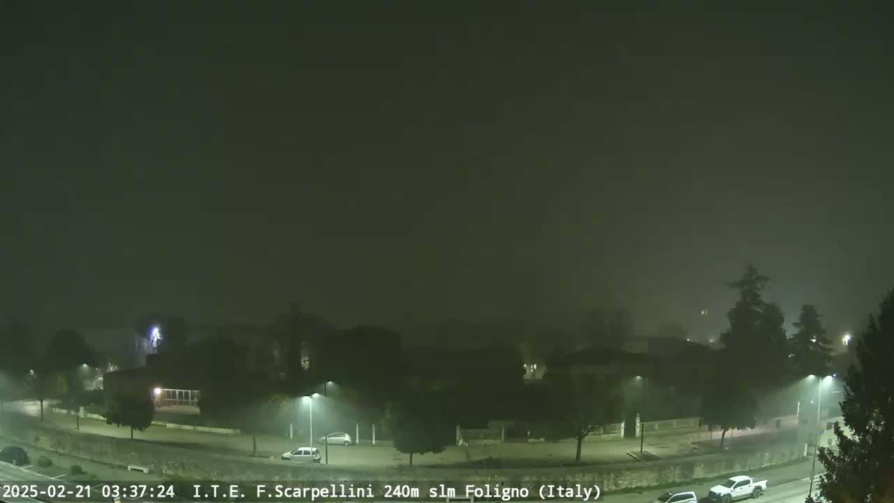 A foggy night scene shows a road curving through a town with sparsely lit buildings and a few vehicles.