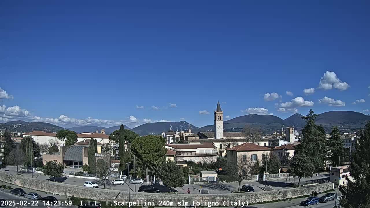 A partly sunny day reveals a town nestled in a valley, with mountains in the background and a tall bell tower prominently featured among the buildings.