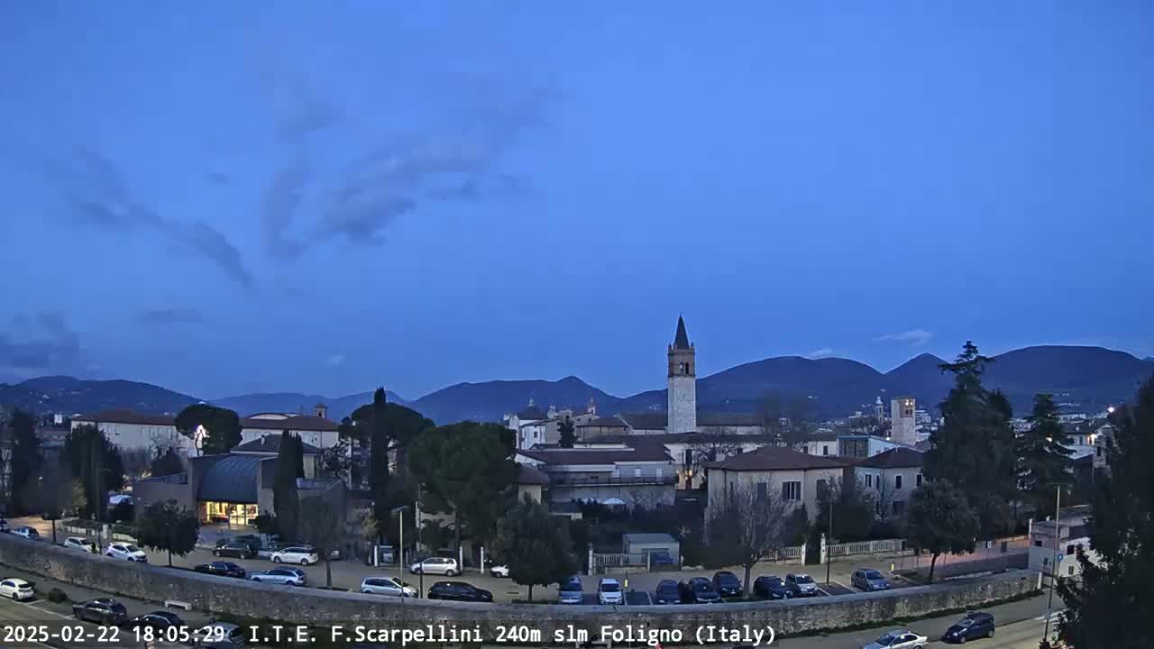 A twilight view of a town nestled in a valley, under a mostly clear blue sky, with mountains in the background.