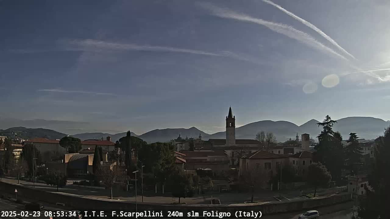 A mostly sunny day reveals a town nestled in a valley, with mountains in the background and contrails streaking across the pale blue sky.