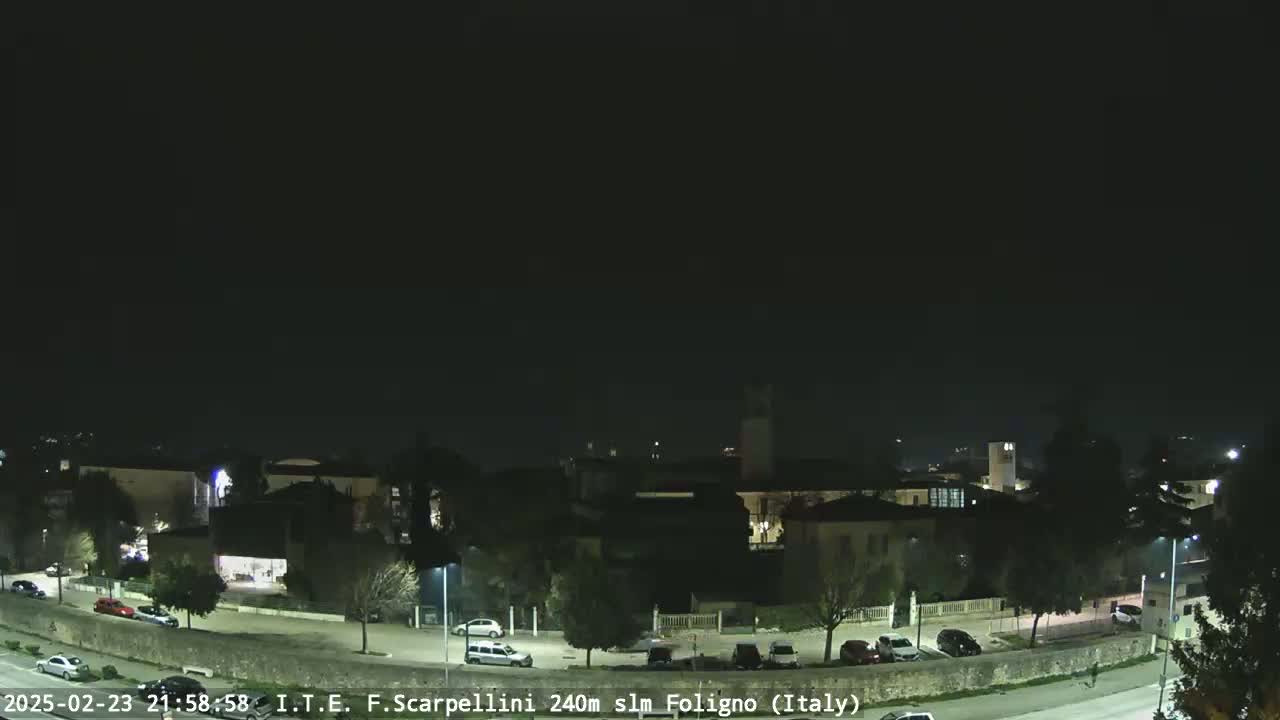 A nighttime, clear-sky view of a town with parked cars in a lot and along a road curving through the scene.
