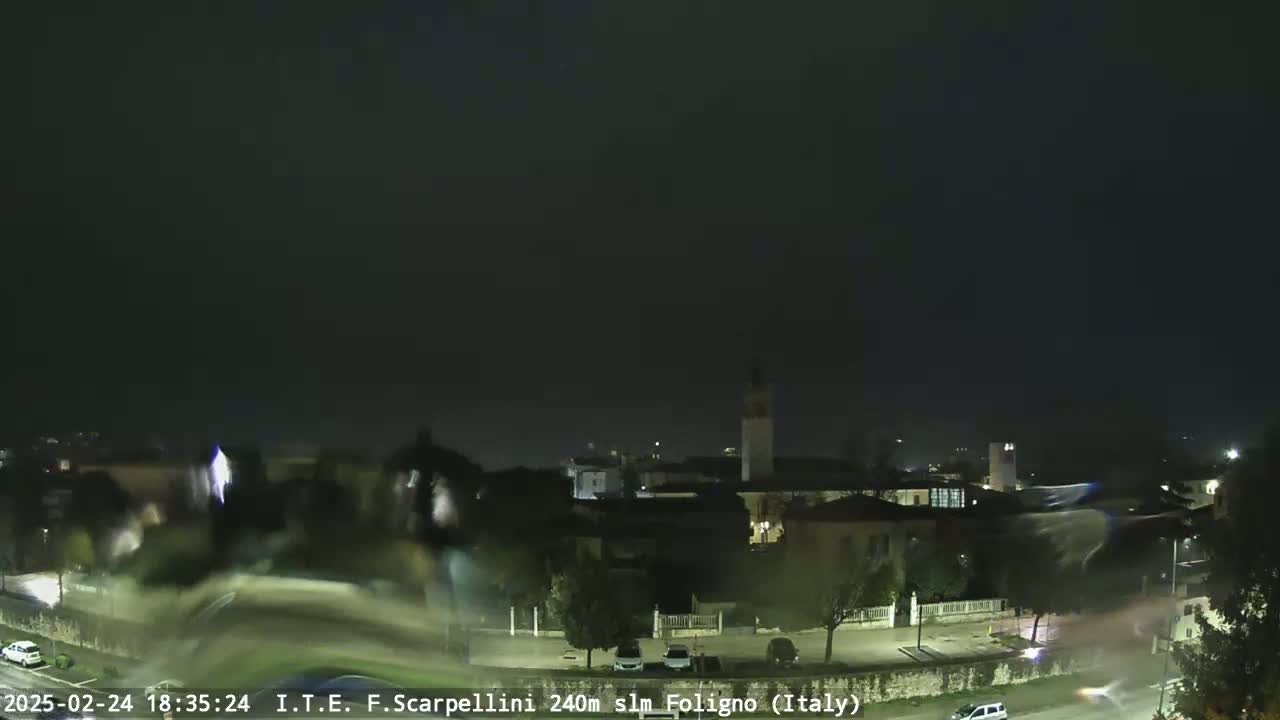 A nighttime view of a town, possibly in a valley, under a dark, overcast sky with some faint light pollution visible in the distance.