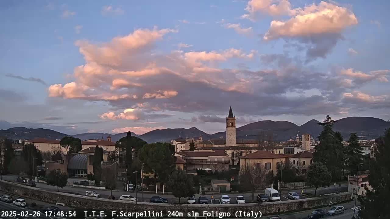A picturesque town nestled in a valley, under a pastel-colored sunset sky with fluffy clouds, is bordered by mountains in the background.