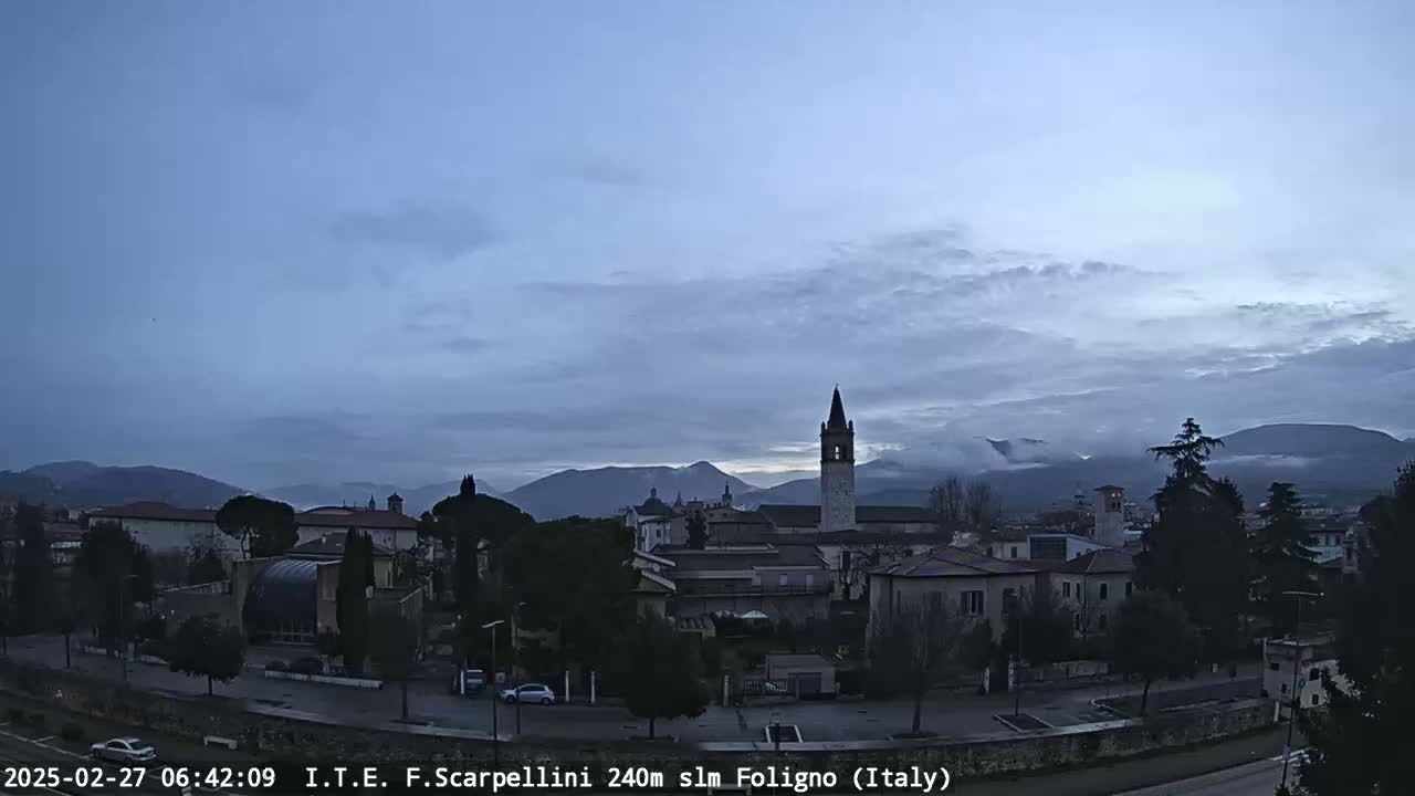 A pre-dawn view of a town nestled in a valley, under a mostly clear, dusky sky with distant mountains shrouded in fog.
