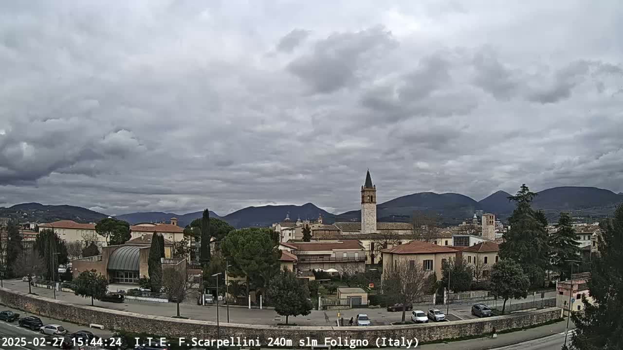A town nestled in a valley under a cloudy sky, with mountains in the background and a tall, slender tower visible in the center.
