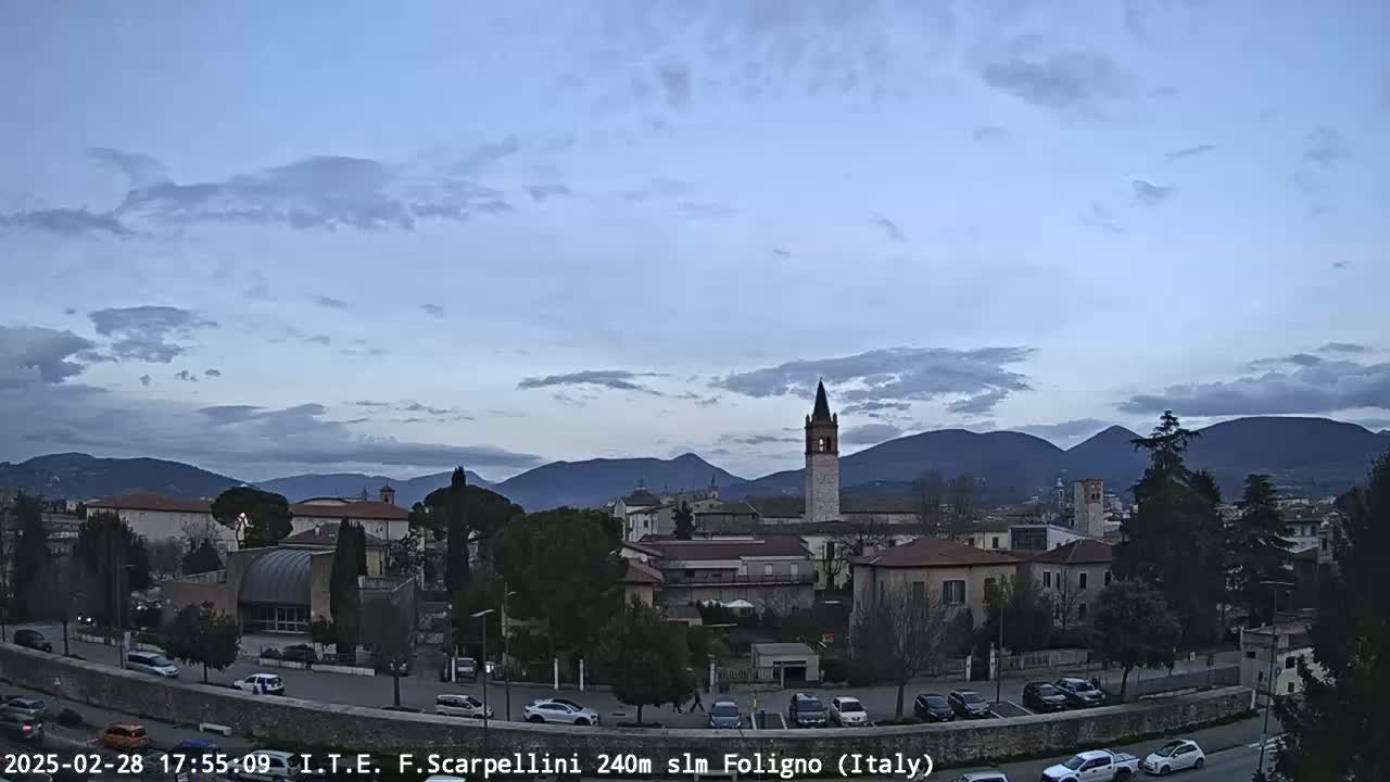 A partly cloudy twilight sky overlooks a town with buildings, cars, and a tall bell tower, backed by distant mountains.