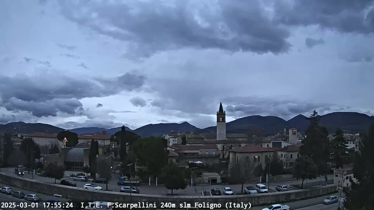 A town with a tall bell tower is nestled in a valley beneath a cloudy, overcast sky, mountains visible in the distance.