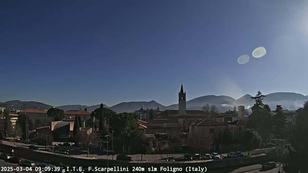 A clear, sunny day reveals a town nestled in a valley, with mountains in the background and a tall church steeple prominent in the foreground.