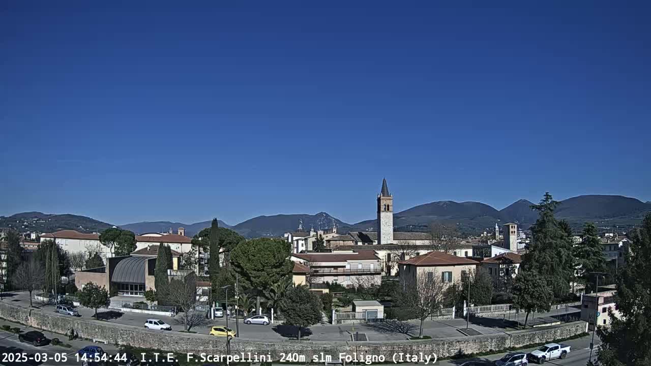 A clear, sunny day reveals a town nestled in a valley, with mountains visible in the background and cars parked along a road in the foreground.