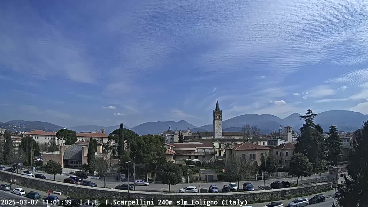 A town with low buildings and a tall bell tower is nestled in a valley under a mostly clear, bright blue sky with thin, wispy clouds and distant mountains.