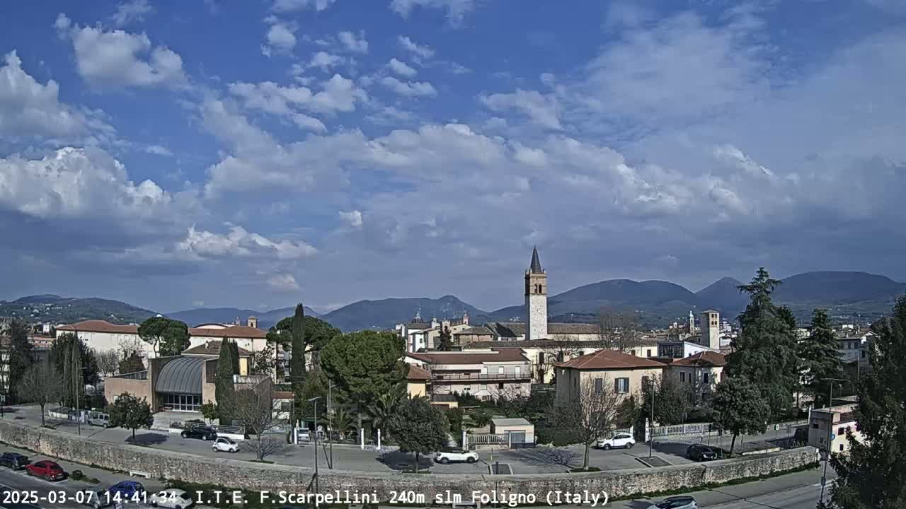A partly cloudy day reveals a picturesque Italian town nestled amongst rolling hills, with cars parked in a lot in the foreground.