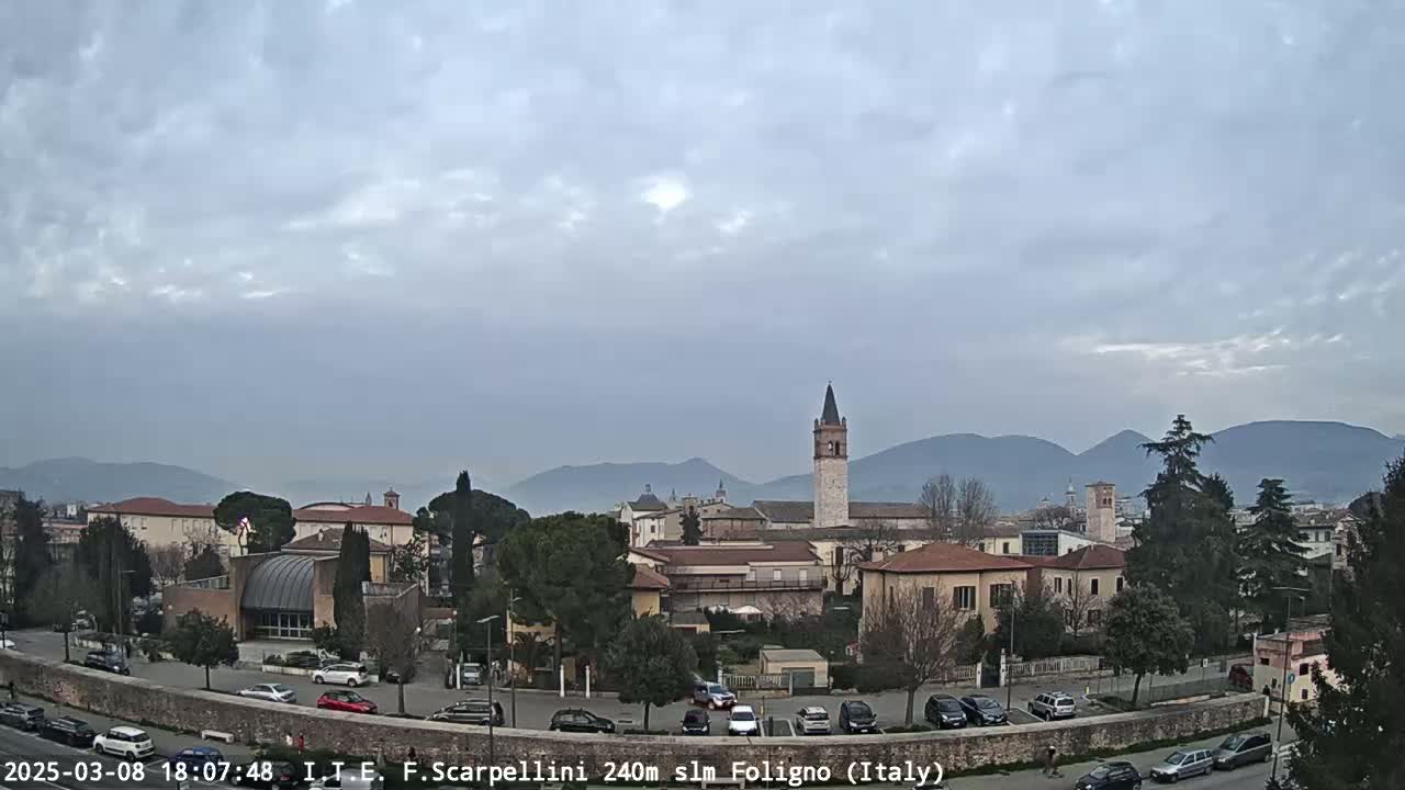 A view of a town with buildings and a tall bell tower, under an overcast sky, with mountains in the background.