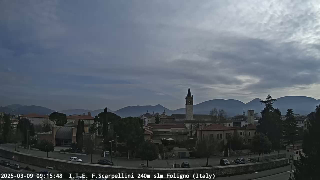 A cloudy day reveals a cityscape with low-rise buildings, a tall bell tower, and distant mountains.