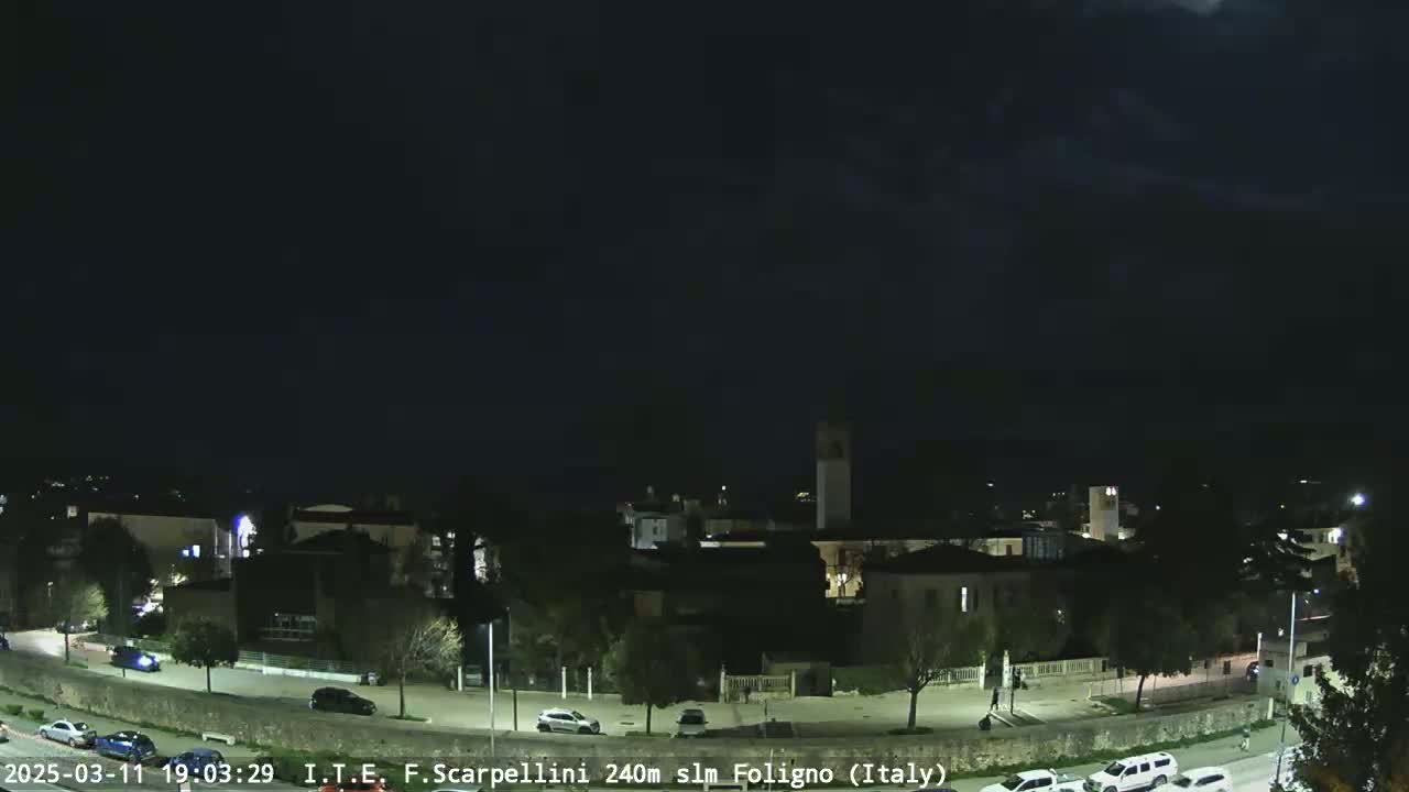 A nighttime view of a town with some cars parked and driving along a road, under a dark, mostly cloudy sky.
