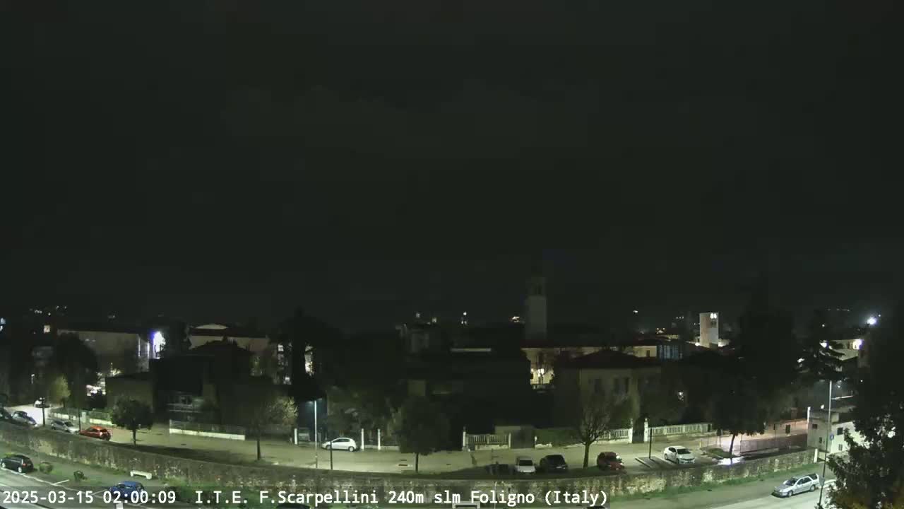 A nighttime, overcast view of a town with sparsely lit streets and parked cars.
