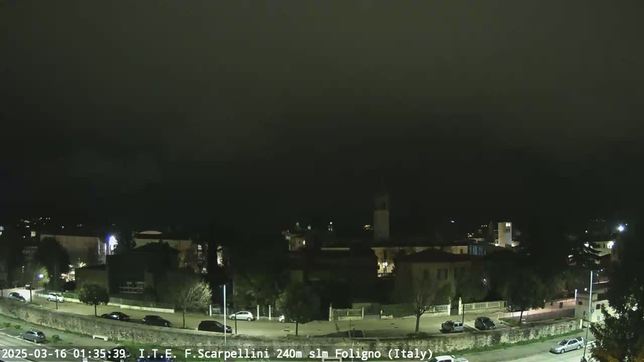 A nighttime view of a town with a few cars parked and driving on roads under a hazy, dark sky.