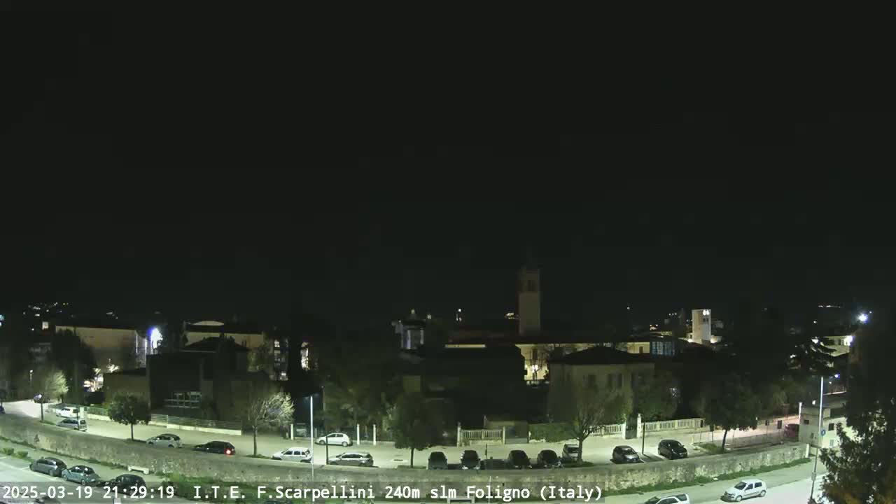 A nighttime view of a town with buildings, streets, and parked cars under a clear, dark sky.