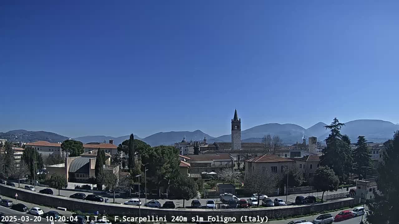 A clear, sunny day reveals a town nestled in a valley, with mountains in the background and a church steeple prominently featured.