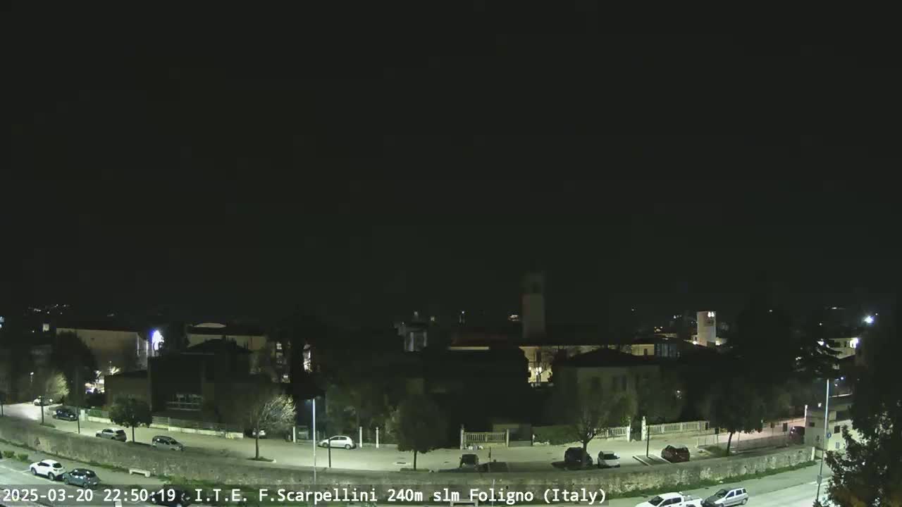 A nighttime, long shot of a town with sparsely lit buildings and a few cars parked along a road.