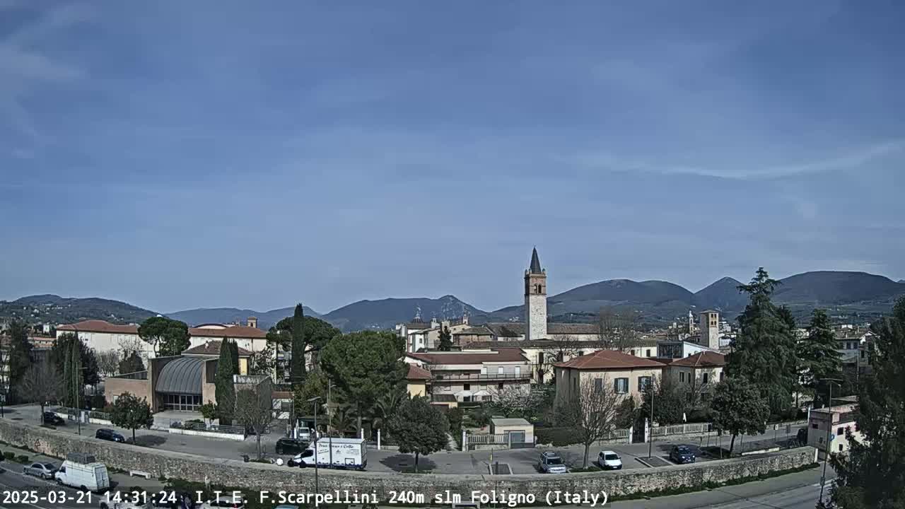 A picturesque Italian town, nestled amongst rolling hills under a clear blue sky, features a prominent bell tower and a variety of buildings.